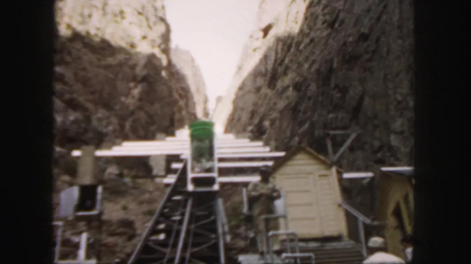 1958 Colorado frame showing visitors or staff near a funicular station with a green car and rugged rock faces behind it.