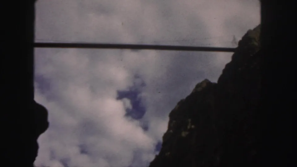 1958 Colorado 8mm frame looking up between dark mountain peaks toward white clouds and blue sky, crossed by a thin cable.