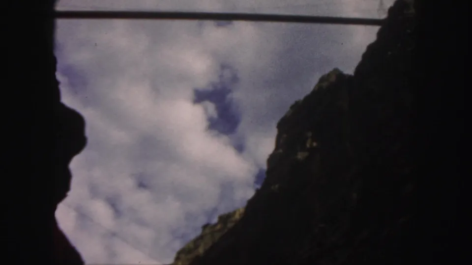 The frame shows a steep shadowed Colorado mountain peak against white clouds and blue sky in grainy 1958 8mm footage.
