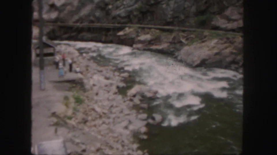 1958 Colorado frame of a whitewater river below rocky slopes, with small visitors visible near the bank.
