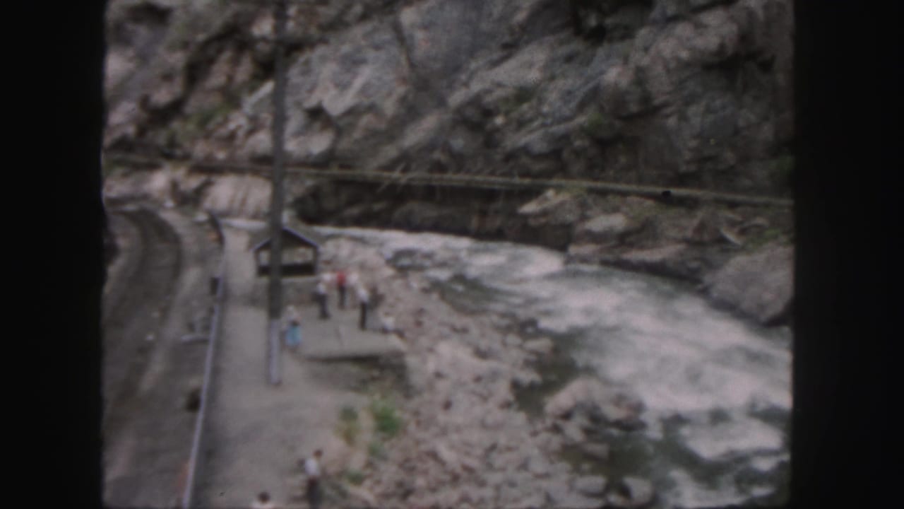 1958 Colorado home-movie frame showing a small riverside platform, people, utility poles, and rocky canyon walls beside fast water.