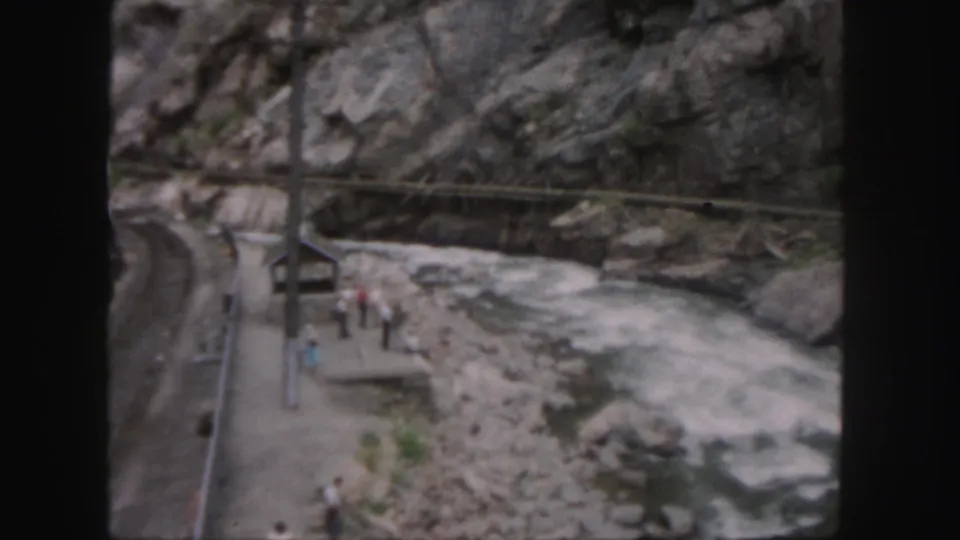 1958 Colorado home-movie frame showing a small riverside platform, people, utility poles, and rocky canyon walls beside fast water.