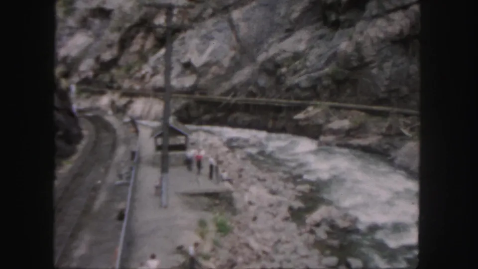 1958 Colorado canyon view with visitors on a narrow platform, a river bend, poles, and rock faces visible from above.