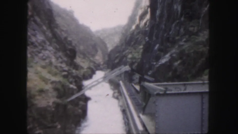 1958 Colorado home-movie frame looking down a narrow rocky canyon with a river, bridge, rails or roadbed, and a roofed structure.