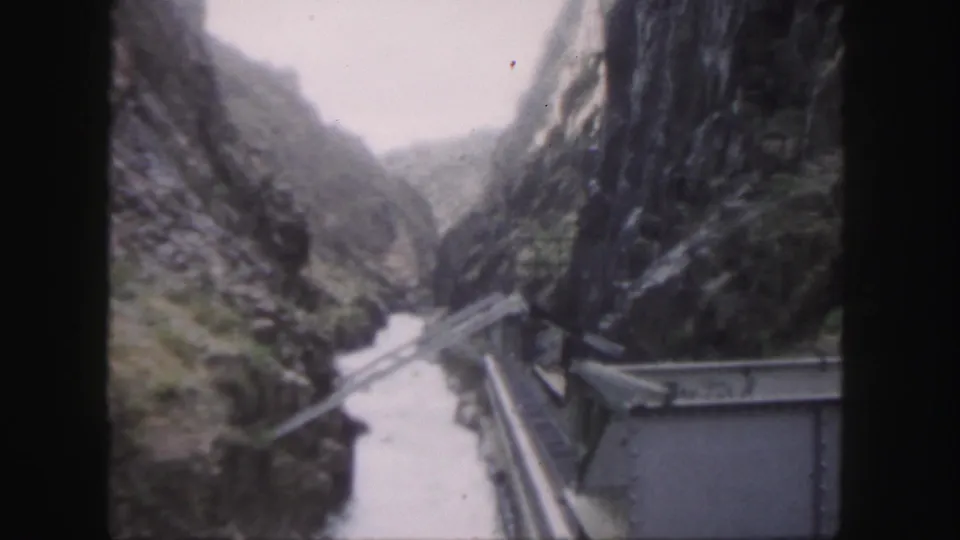 1958 Colorado canyon scene showing a river below, a bridge crossing, steep rock walls, and a nearby roofed rail or utility structure.