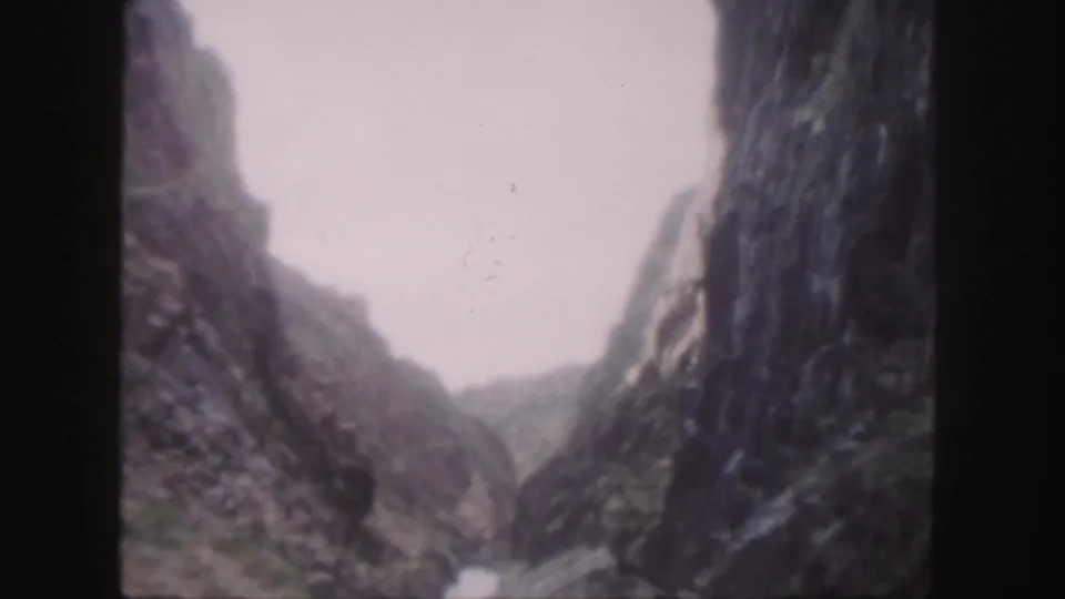 1958 Colorado 8mm frame of a rocky mountain river corridor, with a cable or narrow bridge structure crossing the scene.