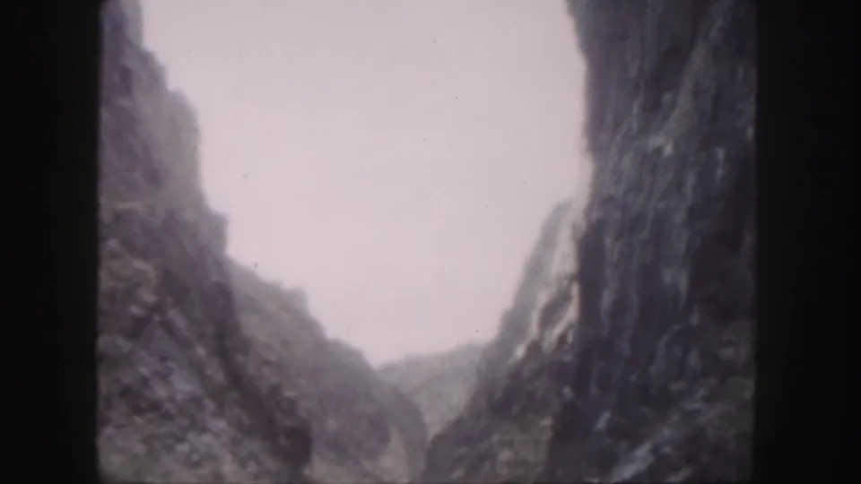 1958 Colorado home-movie frame showing a narrow mountain river valley framed by tall rock walls and hazy distant terrain.