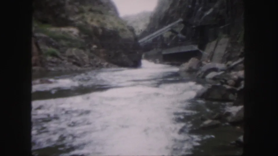 1958 Colorado 8mm frame showing a river below a box-like carrier or platform suspended near rough scaffolding in a mountain canyon.