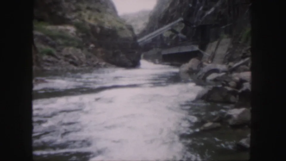 Vintage 8mm view of a Colorado canyon river, with rapids in the foreground and a narrow bridge crossing near the rocks.