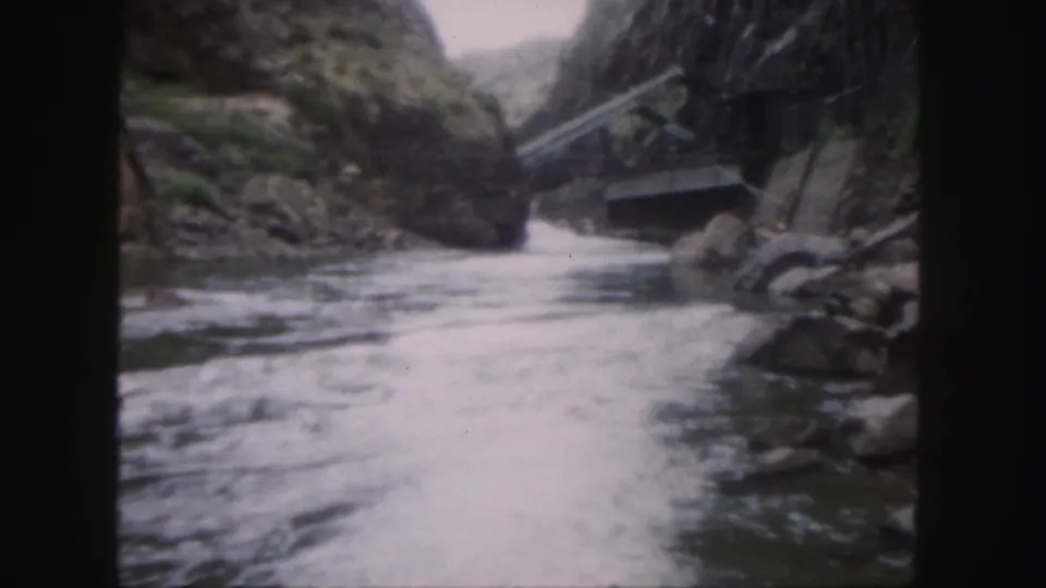 1958 home-movie frame of foaming river water in a Colorado canyon, with rocks and an old bridge partly visible to the right.
