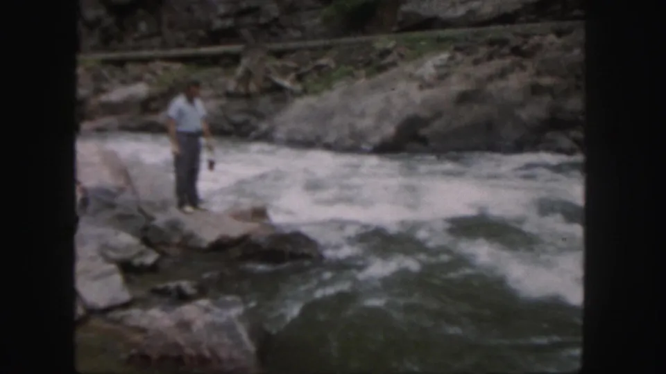 1958 Colorado 8mm frame showing a man in a light shirt and dark pants standing on rocks near fast-moving river water.