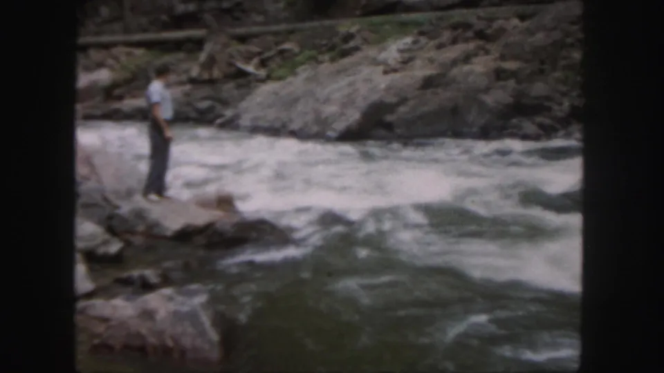 1958 Colorado home-movie frame of a man on rocks beside a turbulent mountain river, surrounded by boulders and canyon walls.