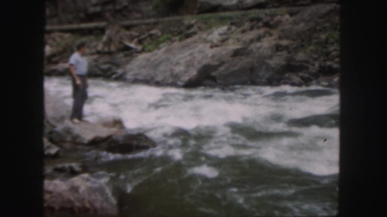 In Colorado 1958 home-movie footage, a man stands on a rocky edge beside churning river currents.