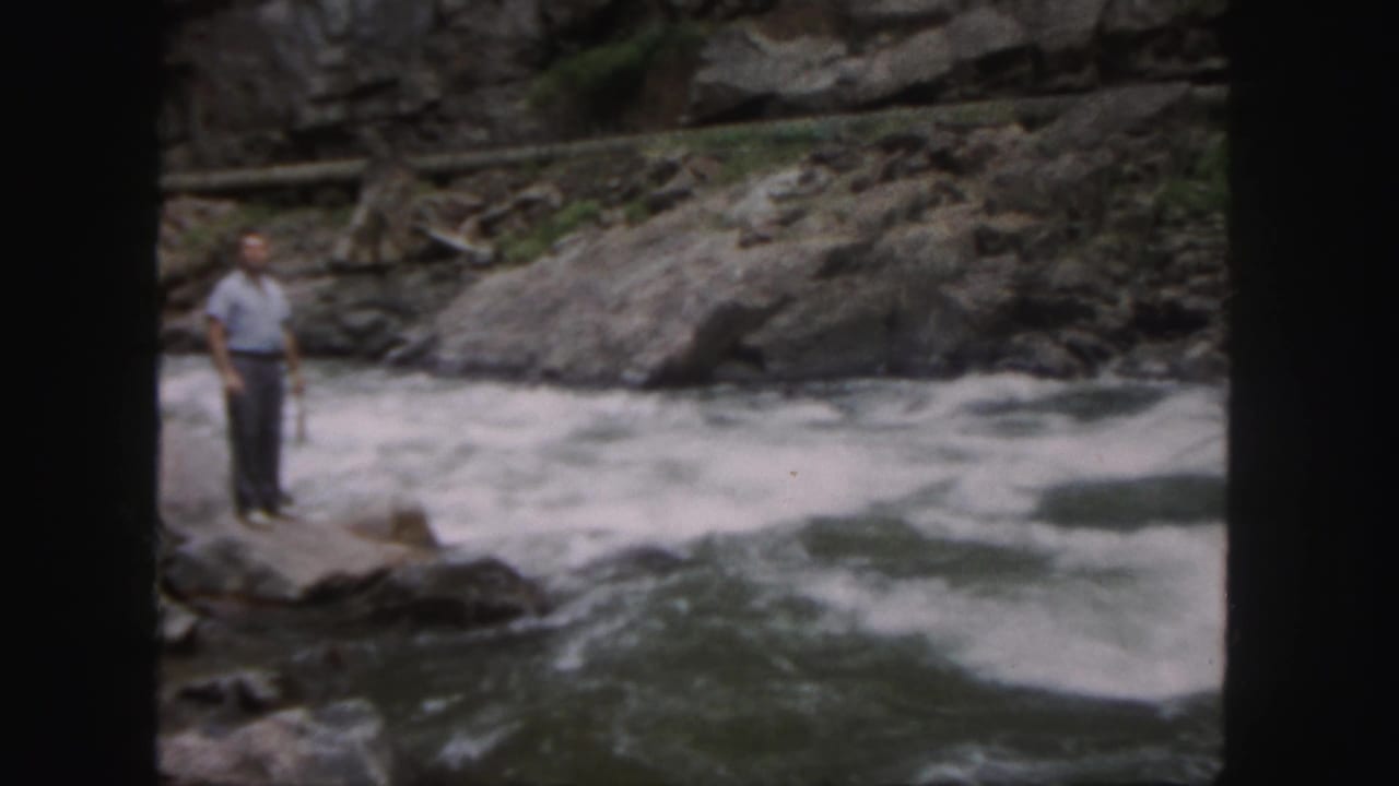Man stands near the edge of rushing water in a rocky river setting.