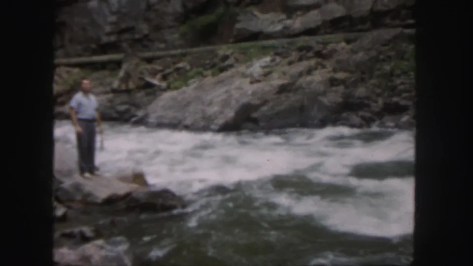The man remains on a rock ledge beside turbulent Colorado river water, with boulders and a rough bank behind him.