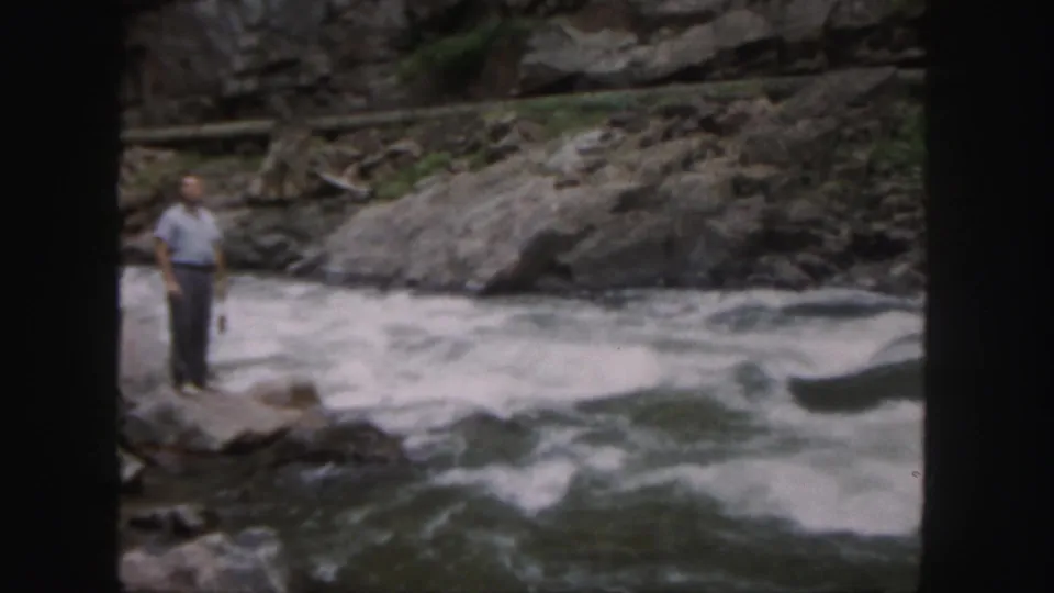 A soft 8mm frame shows the man beside whitewater and dark current, with rocks and a canyon-like bank in the background.