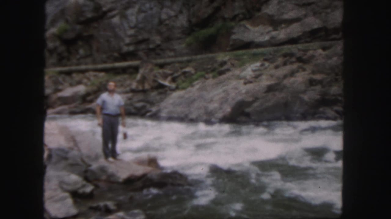 Man standing on rocks beside a fast whitewater river.