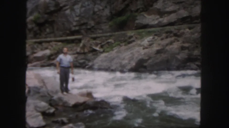 In 1958 Colorado home-movie footage, a man stands on a rocky bank beside rushing whitewater in a canyon-like setting.