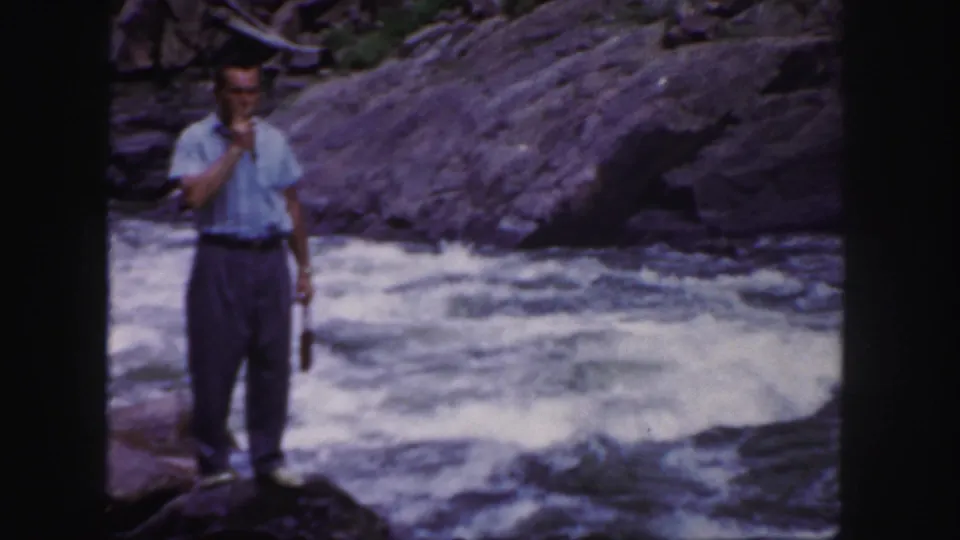 The man faces the camera area while smoking on a rock ledge, with foaming river water and dark stone behind him.
