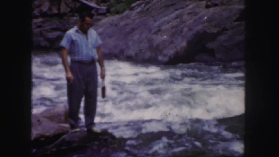 A man stands on riverside rocks in Colorado, looking down toward the rushing water with a small bottle hanging from one hand.