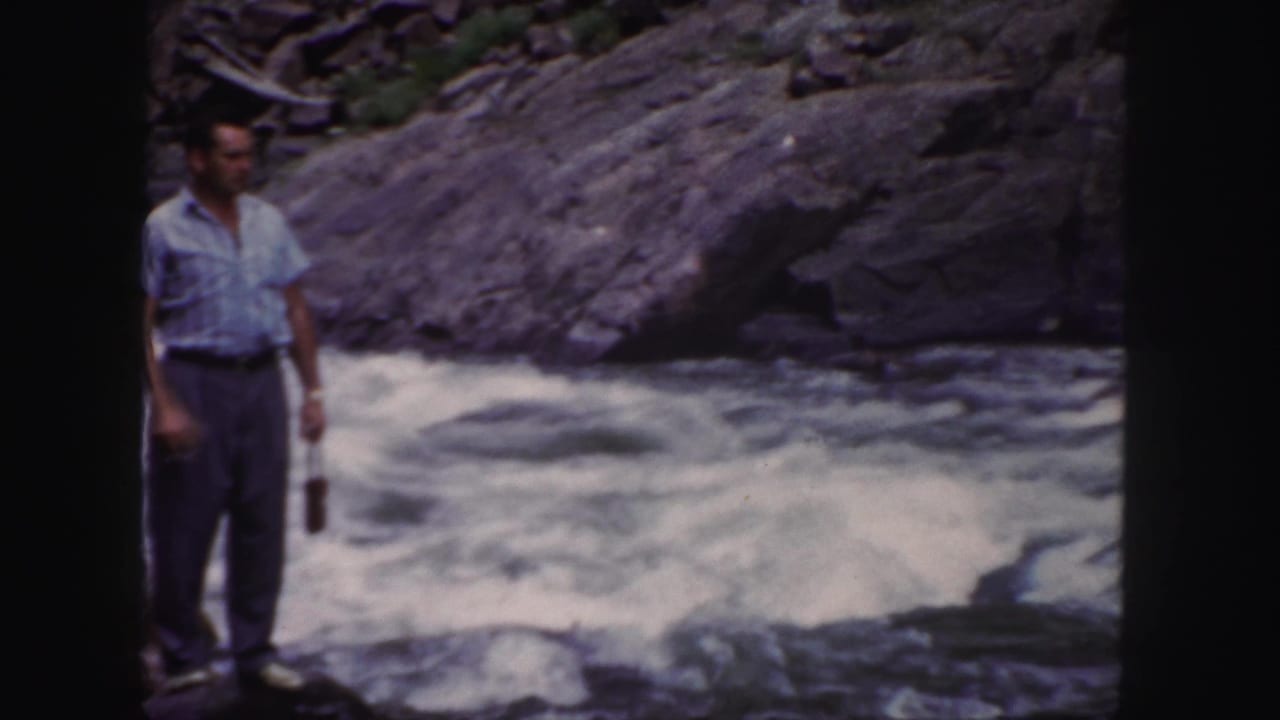 The man remains near the Colorado river, holding a small object while whitewater moves behind him.