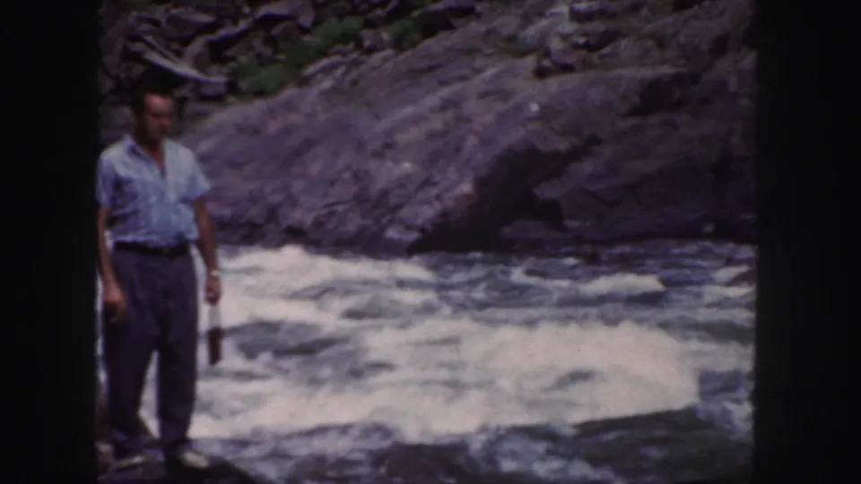 A handheld 8mm frame shows the man near the left edge, with foaming river water and a rocky bank behind him.