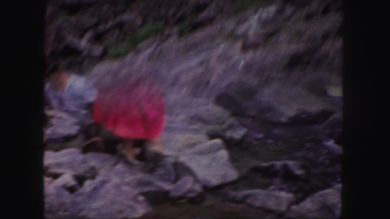 1958 Colorado 8mm frame showing two adults near large boulders and moving water, with the woman blurred in pink as she crosses rocks.