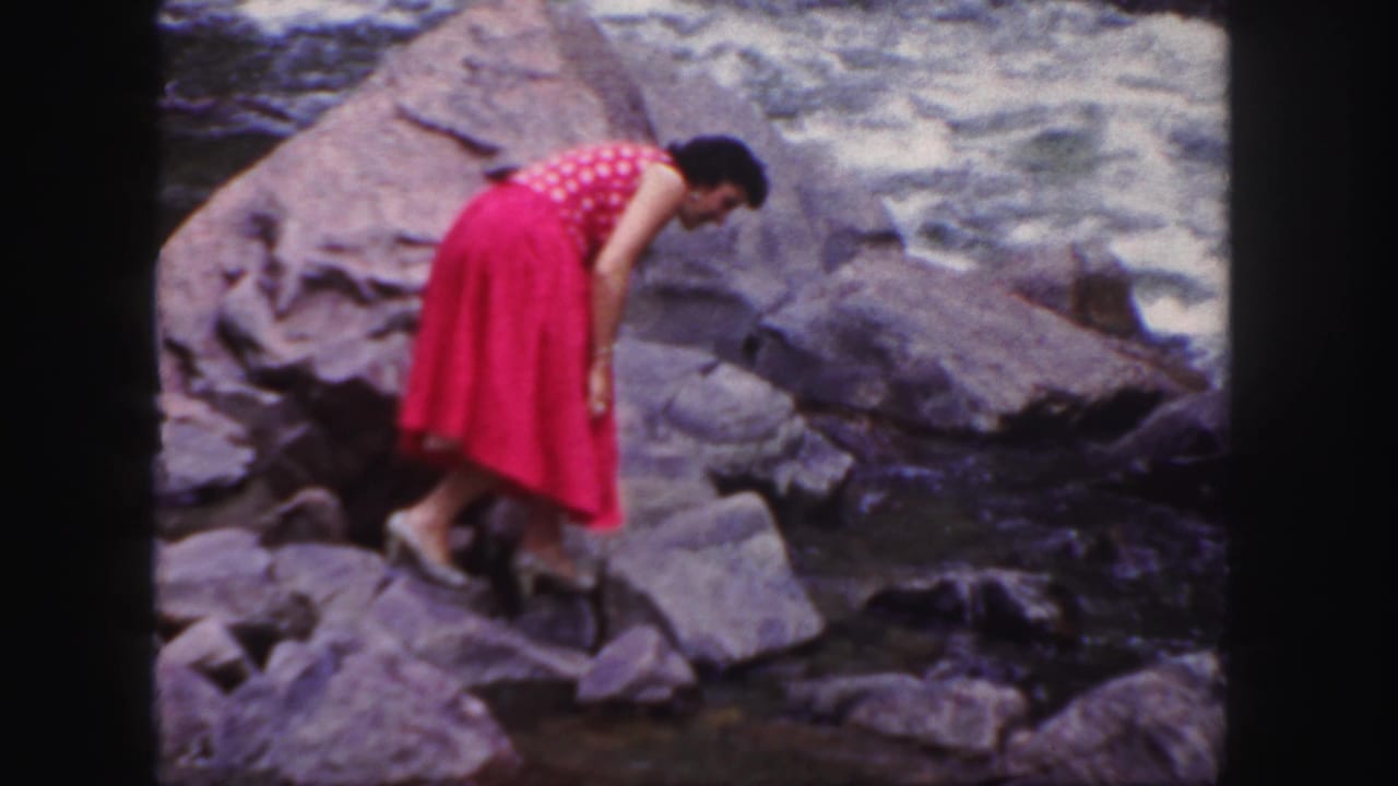 Woman in a pink polka-dot dress bends over rocks beside rushing water.