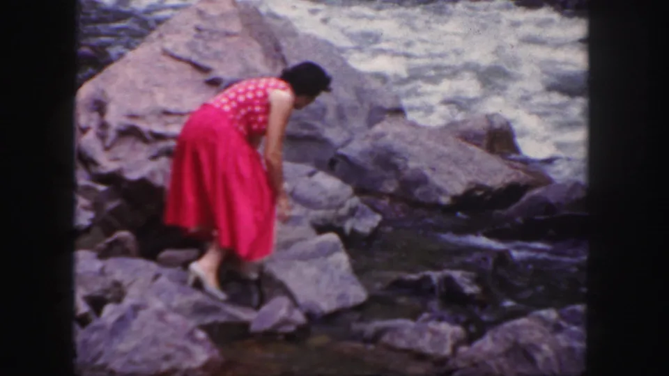 1958 Colorado 8mm frame showing a woman in a bright pink dress bending low on rocks beside a flowing river.