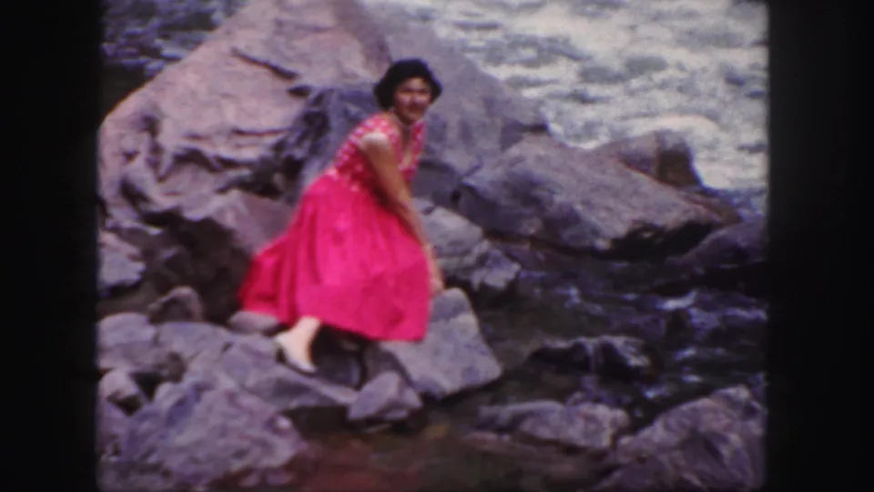 1958 Colorado home-movie frame with a woman in a pink polka-dot dress seated on a riverside rock, smiling back at the camera.