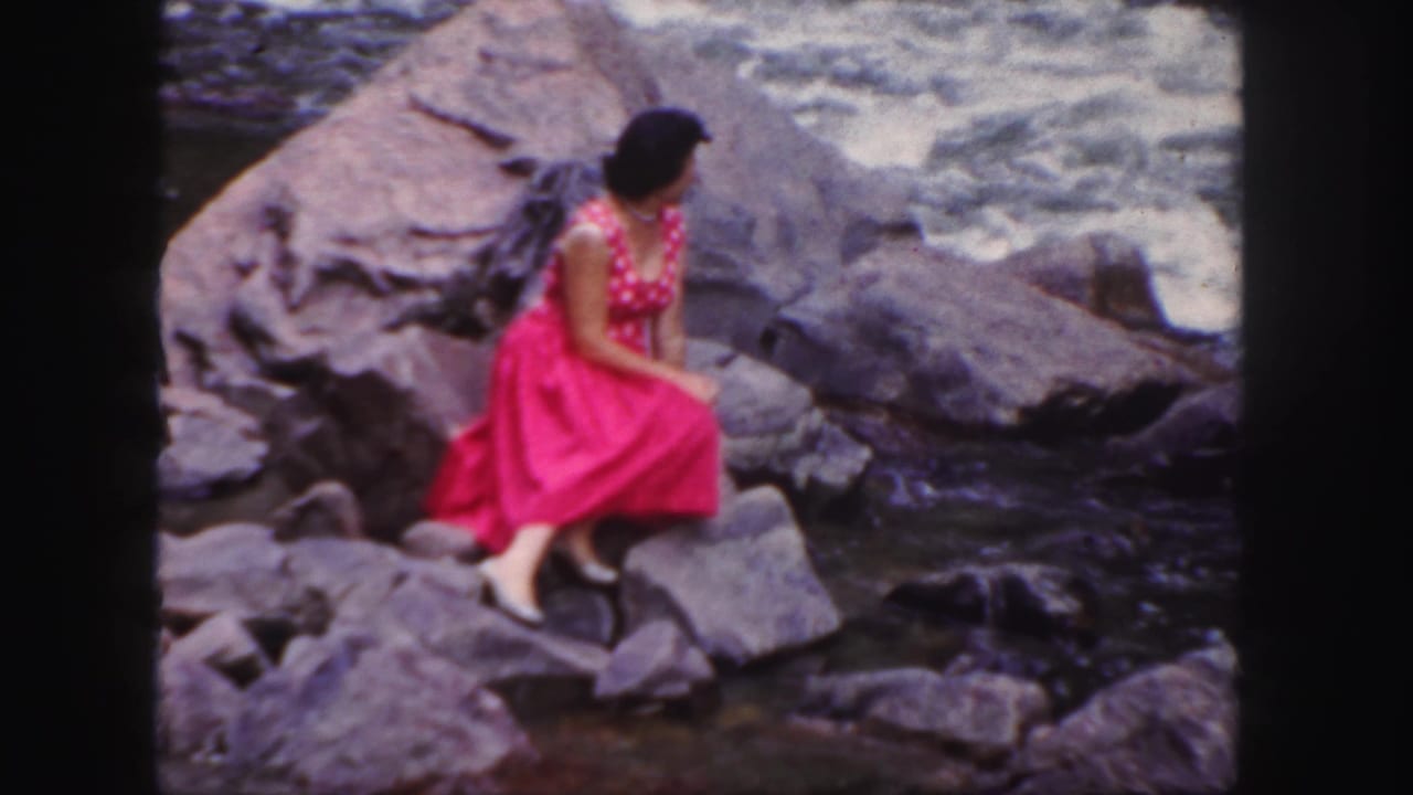 Woman in a bright pink dress sits on rocks beside rushing river water.