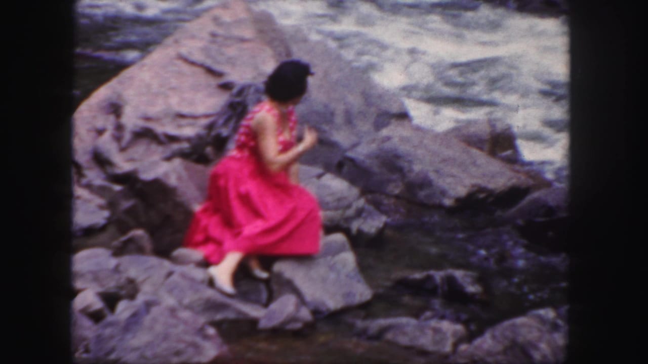 Woman in a pink dress turns while seated on rocks near rushing water.