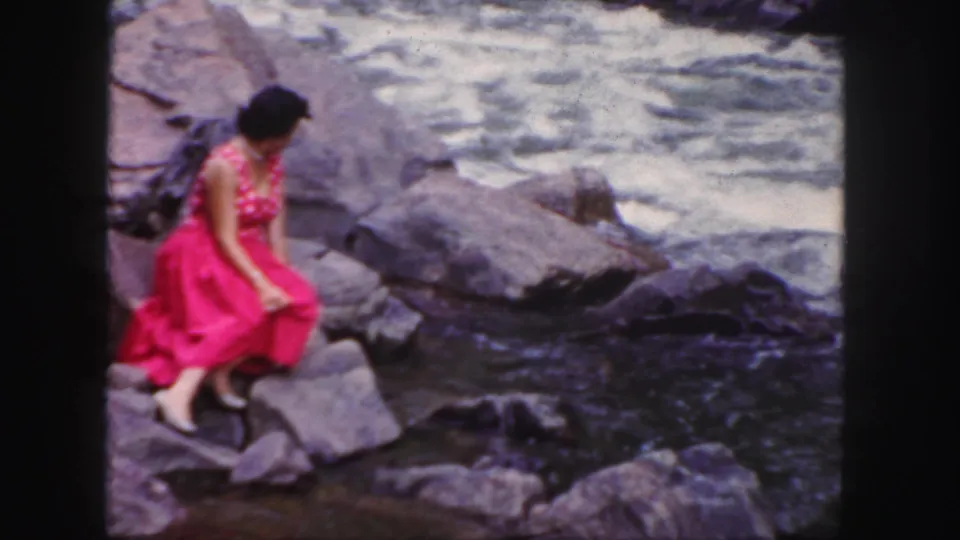 A woman in a bright pink dress sits lower on the rocks, looking downward near dark water and gray stones in this 1958 home movie.