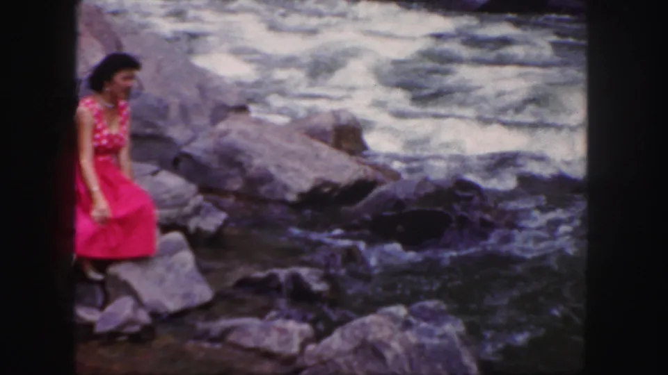 The woman sits near the edge of the rocks, with churning river water filling the background and soft 8mm texture visible.