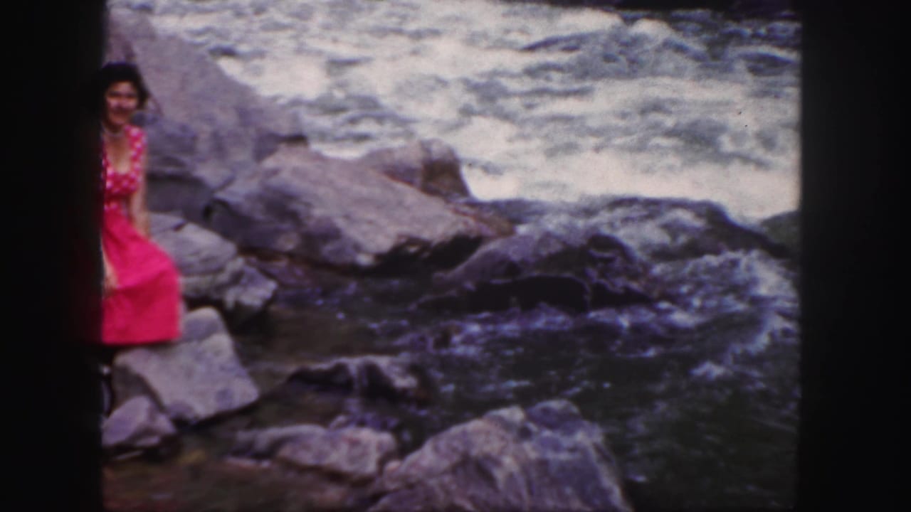 Young woman in a red dress sitting on rocks beside a rushing river.