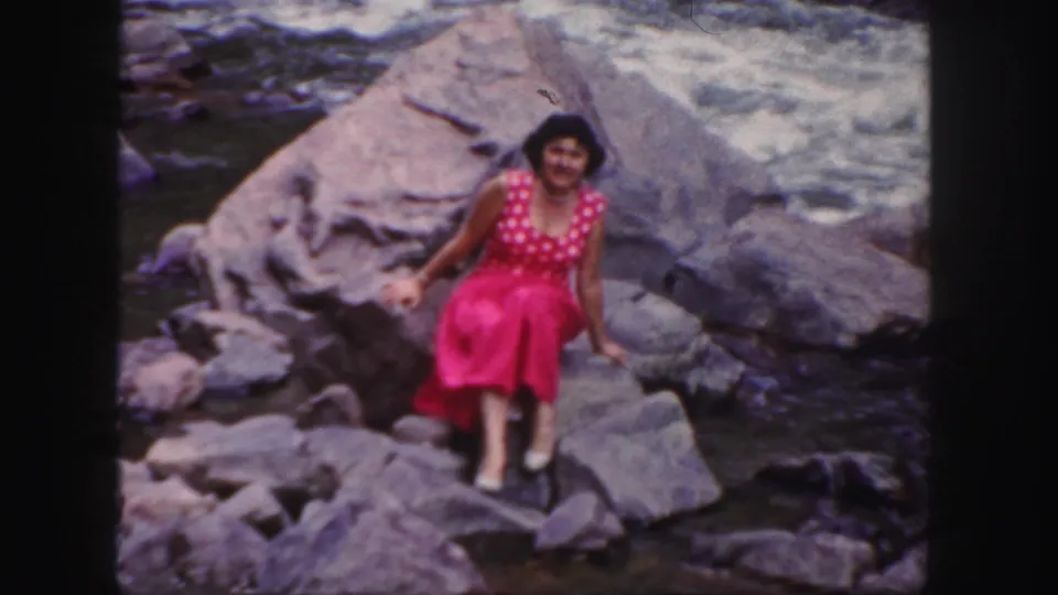 In 1958 Colorado home-movie footage, a young woman in a red polka-dot dress poses on riverside rocks near fast water.