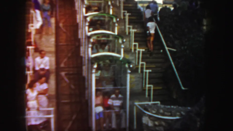 1958 Colorado home-movie frame showing visitors near a steep stairway and a wheel-like rail or lift structure, with faces indistinct.