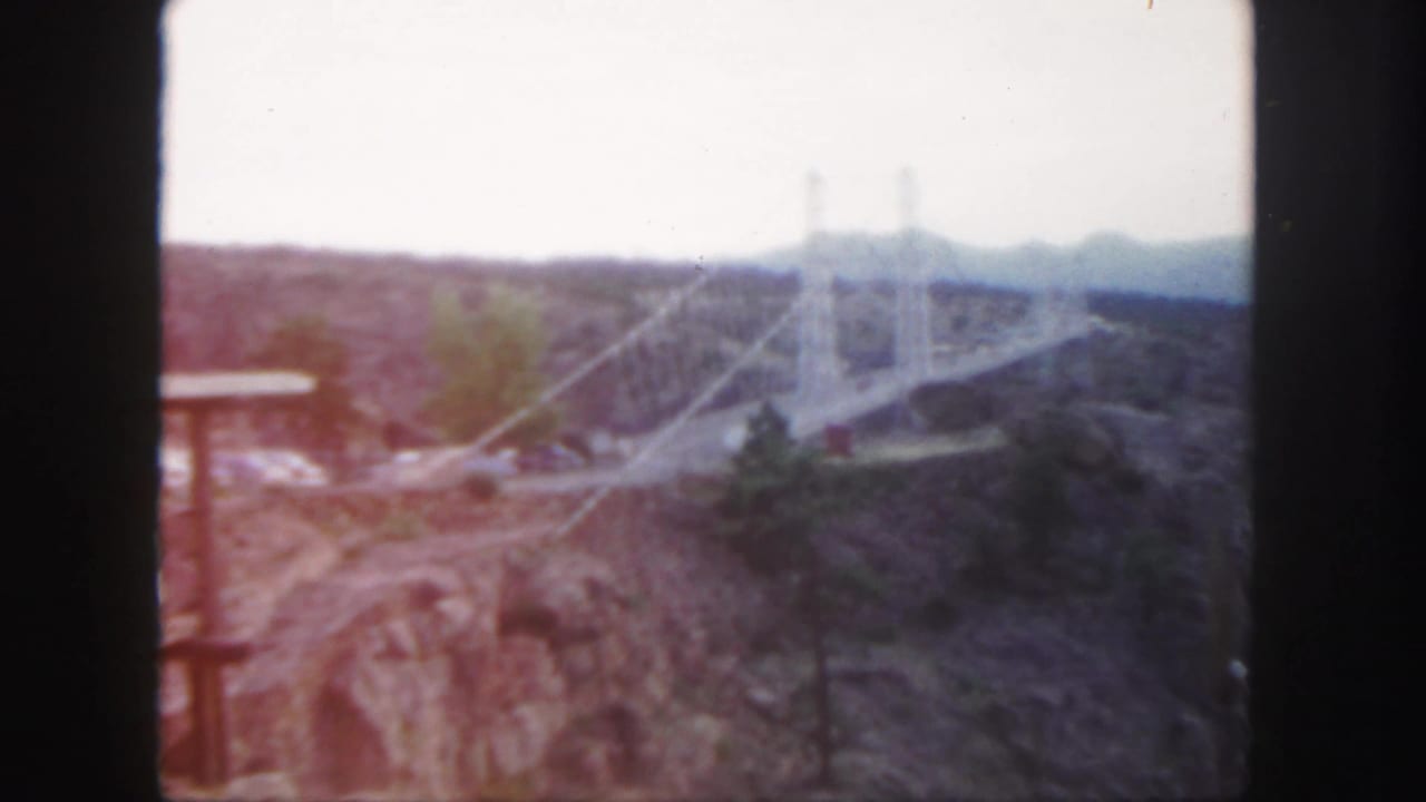 1958 Colorado 8mm frame showing a large white suspension bridge crossing a rugged canyon under a hazy sky.