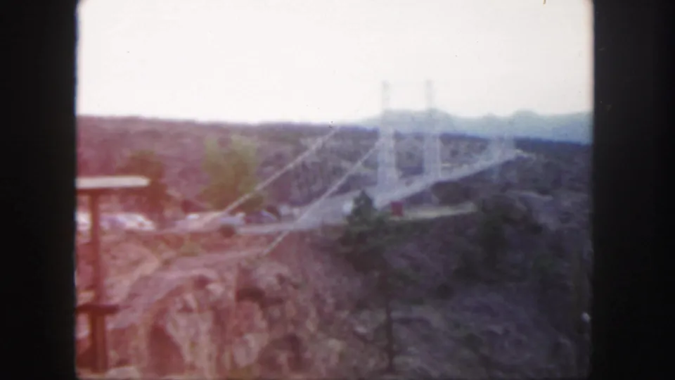 1958 Colorado 8mm frame showing a large white suspension bridge crossing a rugged canyon under a hazy sky.