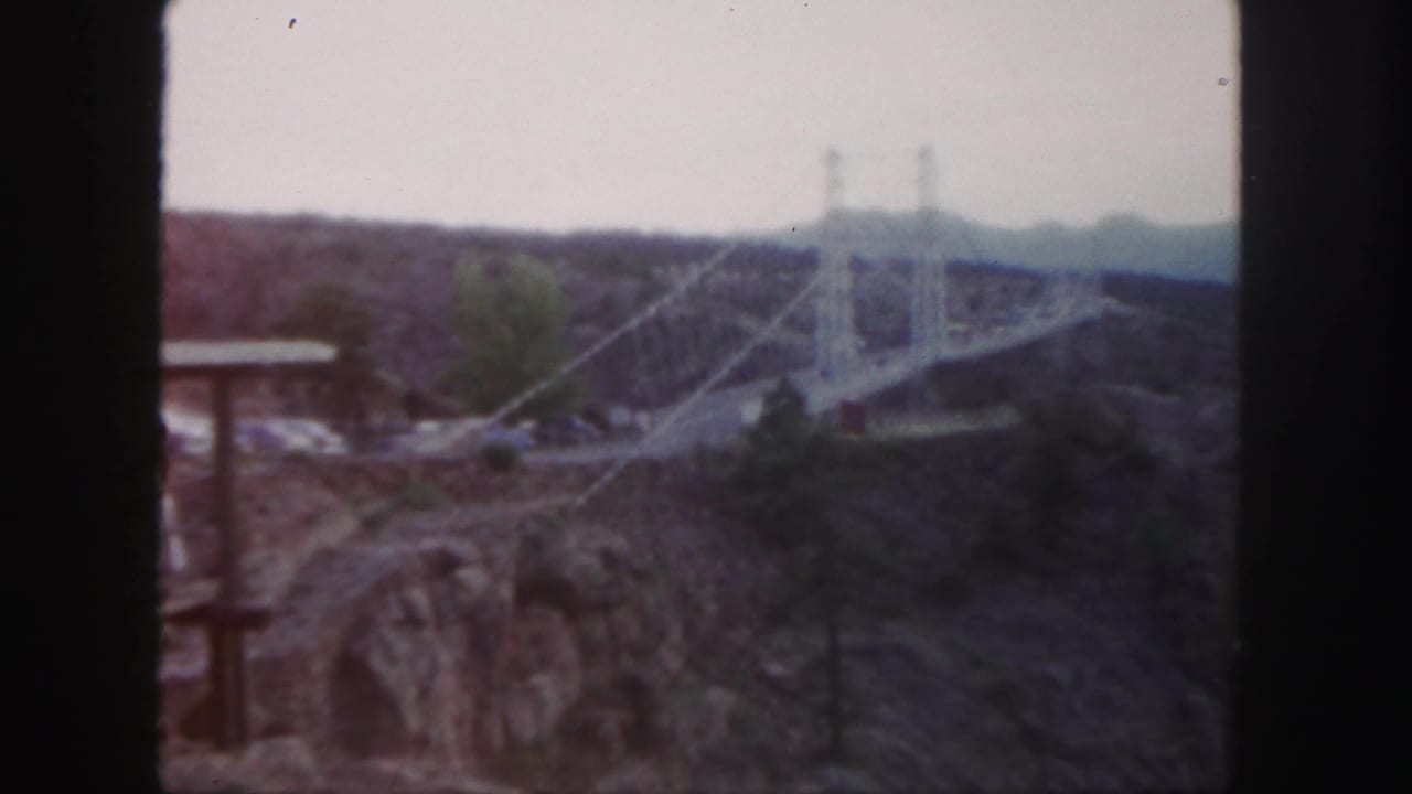 Suspension bridge towers and cables over a canyon with rocky foreground and trees.