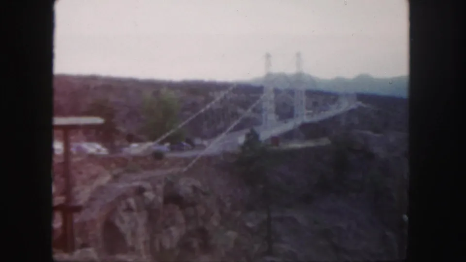 A soft 1958 Colorado 8mm view of a suspension bridge crossing rugged canyon terrain with sparse trees nearby.