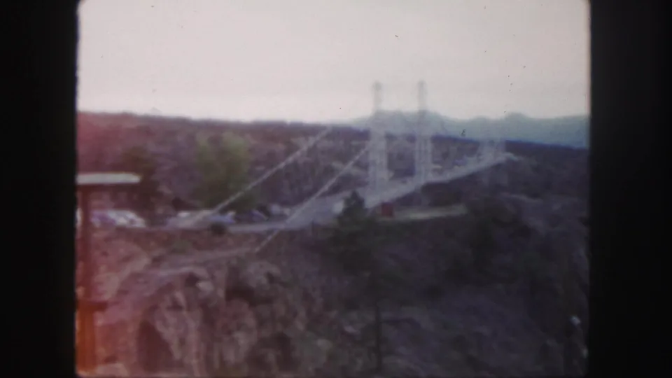 1958 Colorado frame showing the white suspension bridge and cable lines above rocky canyon slopes in faded 8mm color.