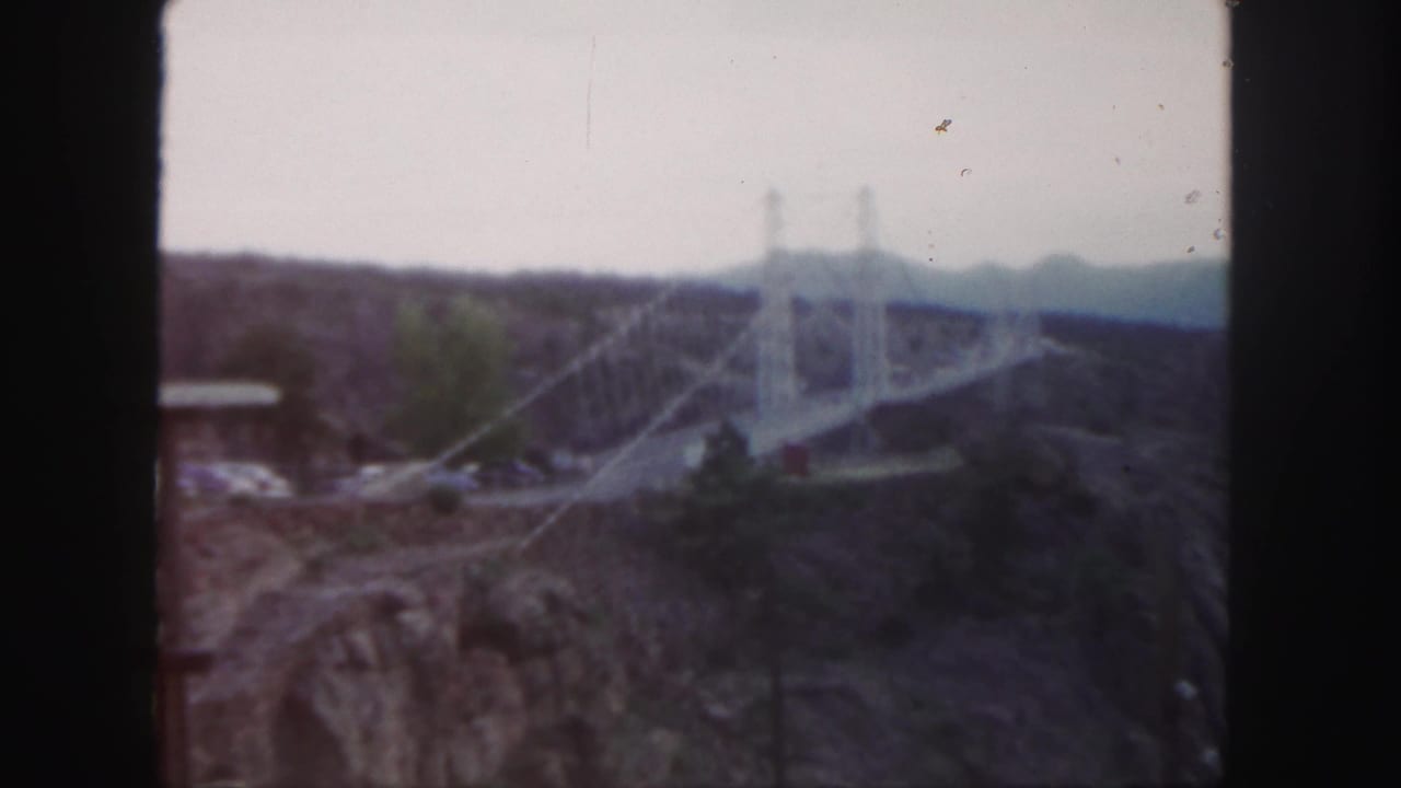 1958 Colorado 8mm frame showing a distant white suspension bridge spanning a rugged canyon with hills beyond.