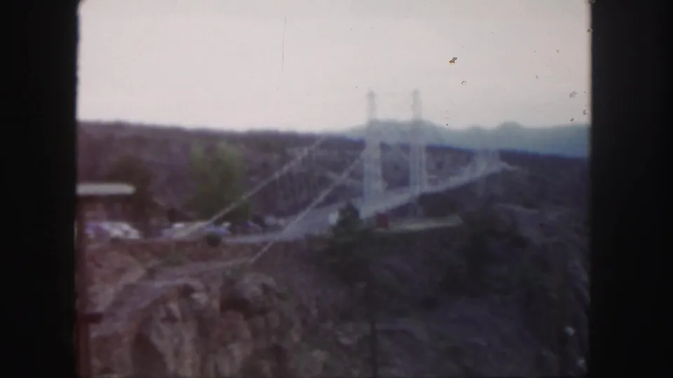 1958 Colorado 8mm frame showing a distant white suspension bridge spanning a rugged canyon with hills beyond.