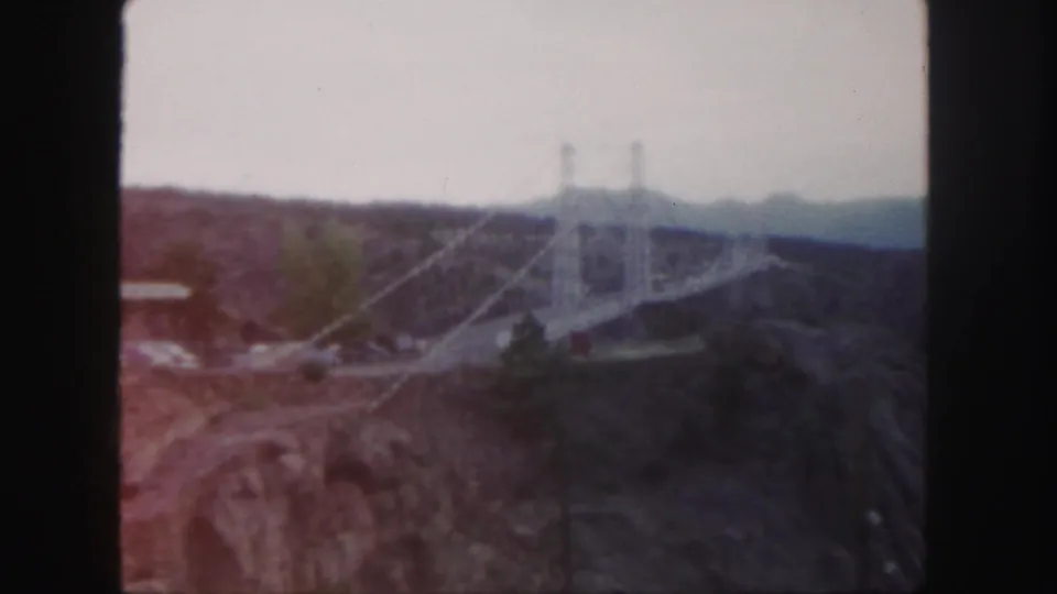 Faded 1958 Colorado home-movie frame with a white suspension bridge, canyon slopes, and distant hills under a light sky.