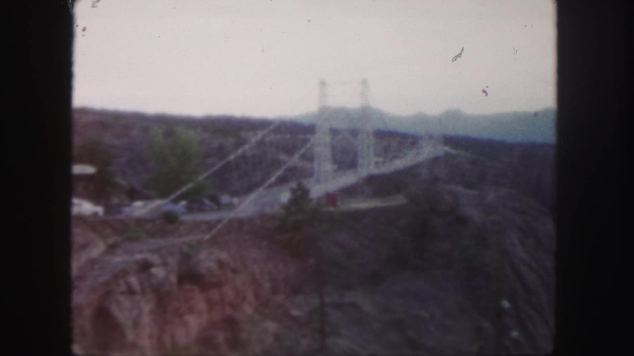 Soft 8mm view of a suspension bridge crossing a rocky canyon with small cars nearby.