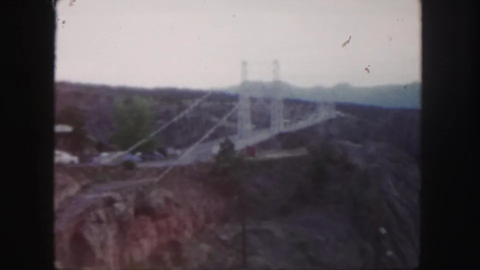 1958 Colorado home-movie frame showing a towered suspension bridge over a canyon, with automobiles and distant mountains visible.