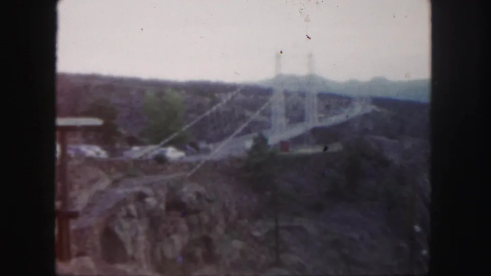 Colorado 1958 8mm frame showing the suspension bridge across rugged canyon terrain with distant hills and visible film grain.