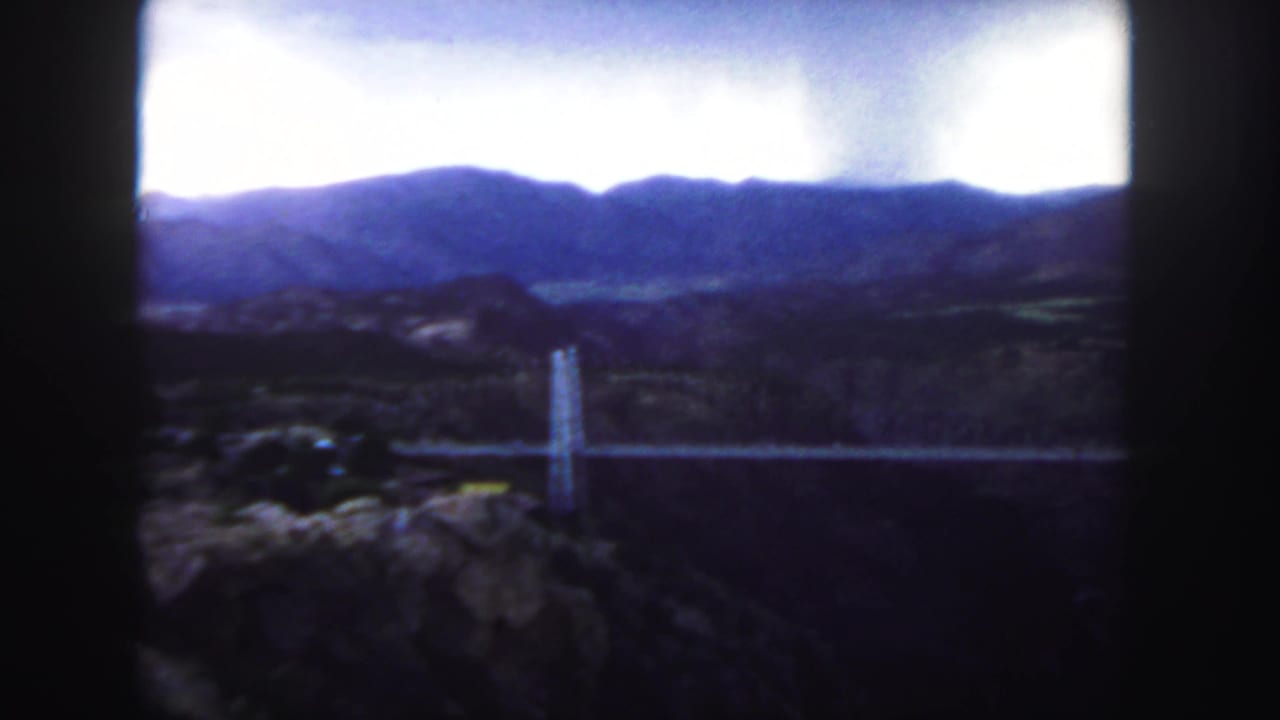 1958 Colorado 8mm frame showing a white suspension bridge crossing a canyon with mountains beyond and dark projector borders.
