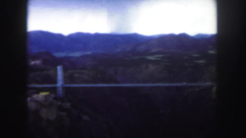 1958 Colorado frame showing the suspension bridge from an overlook, with softened focus, foliage, canyon, and mountains.